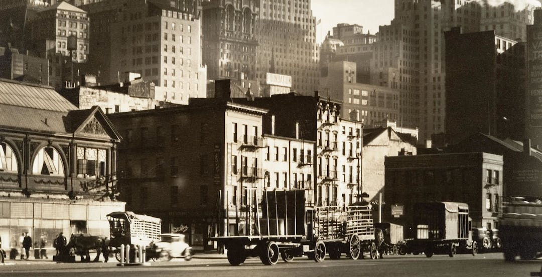 Sepia-tone photograph of old-timey wagon trucks and cars traveling up and down West Street in New York City set against a backdrop of multi-story apartments and midsize skyscrapers.